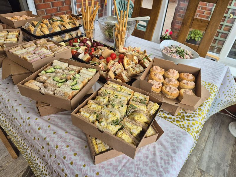 A buffet table laid with sandwich boxes, sausage rolls, a charcuterie platter and quiche