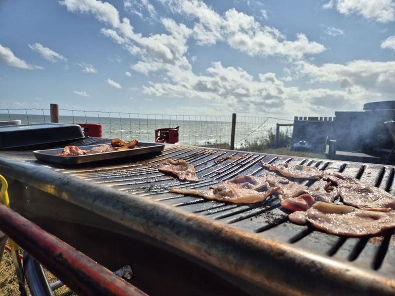 Meat cooking on a large outdoor grill with the sea and blue sky behind
