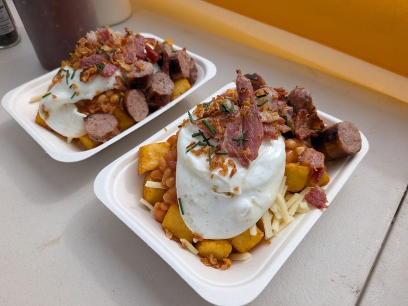 Two trays of cubed potatoes topped with beans, sausage, bacon, cheese and a fried egg